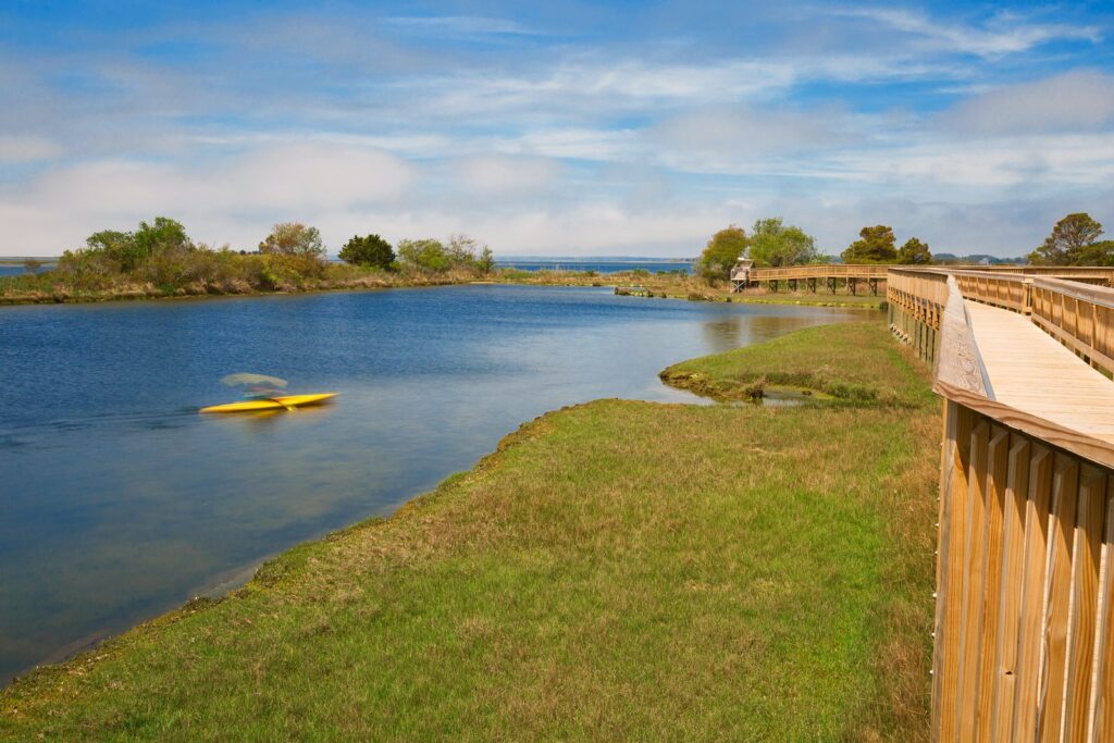 A wooden boardwalk trail beside a calm bay marsh, with a yellow kayak on the water