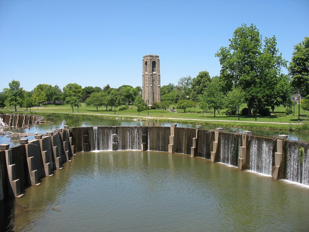 A stone carillon tower beyond a lake and a curved concrete dam with water spilling over, under a blue sky