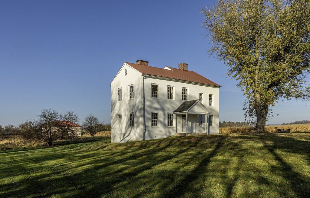 A white two-story farmhouse on a grassy rise with a large tree and long afternoon shadows