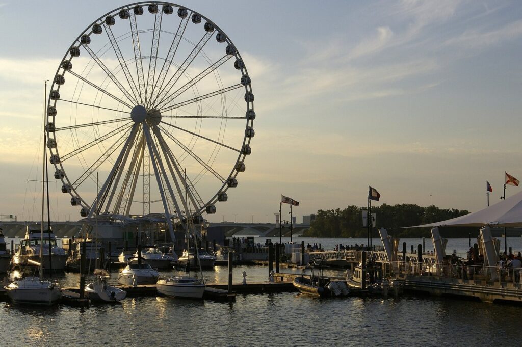 Large white observation wheel beside a marina full of small boats at sunset, with flags flying on the pier