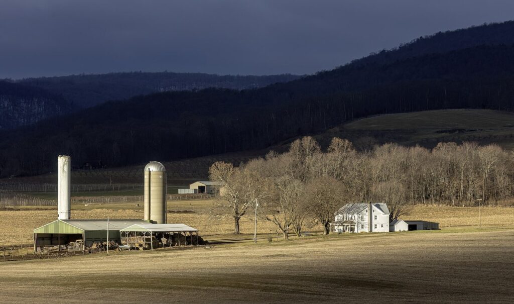 Silos and a white farmhouse on open fields below a long, forested mountain ridge under dark clouds