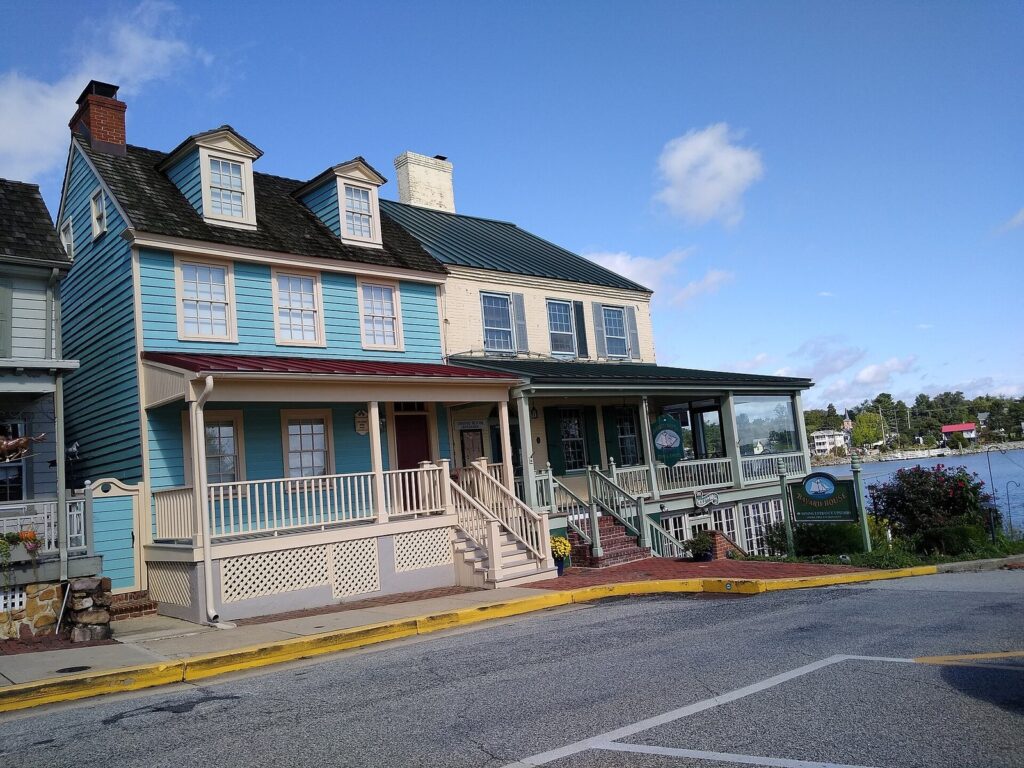 A blue clapboard house with dormer windows and a front porch beside other historic buildings, with water beyond