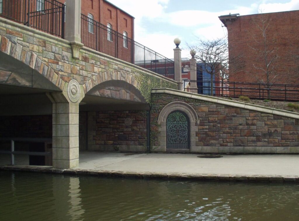 A painted concrete bridge over a creek made to look like carved stone, with brick buildings behind