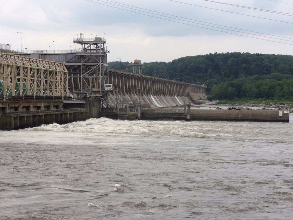 White water churning below the powerhouse and spillway of a long concrete dam, with wooded hills behind