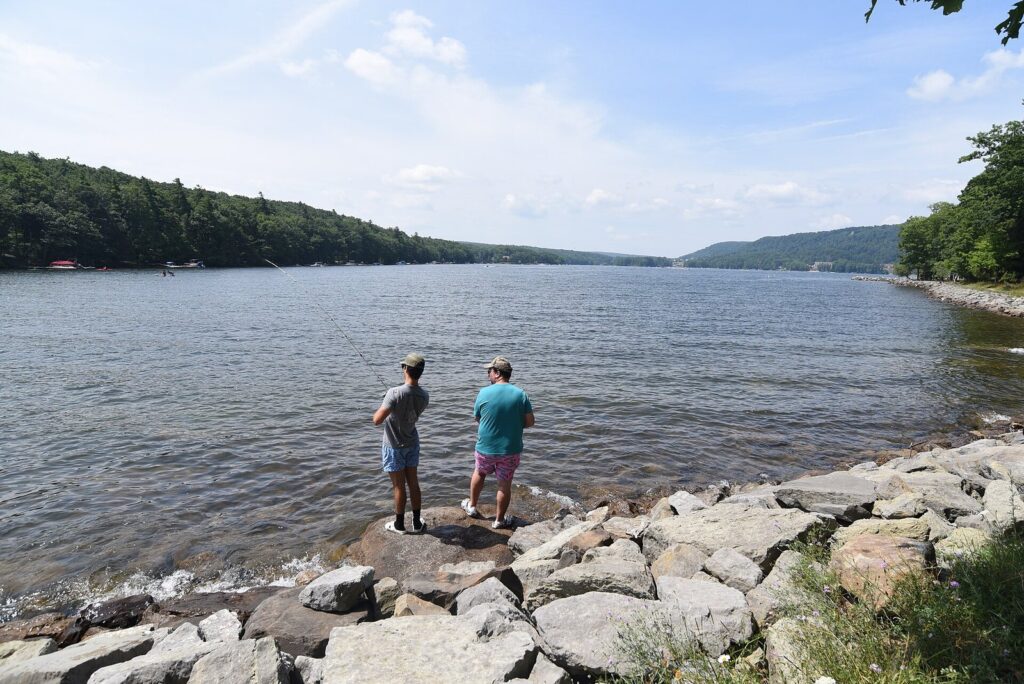 Two people fishing from a rocky point on a summer day, with the lake stretching away between wooded hills