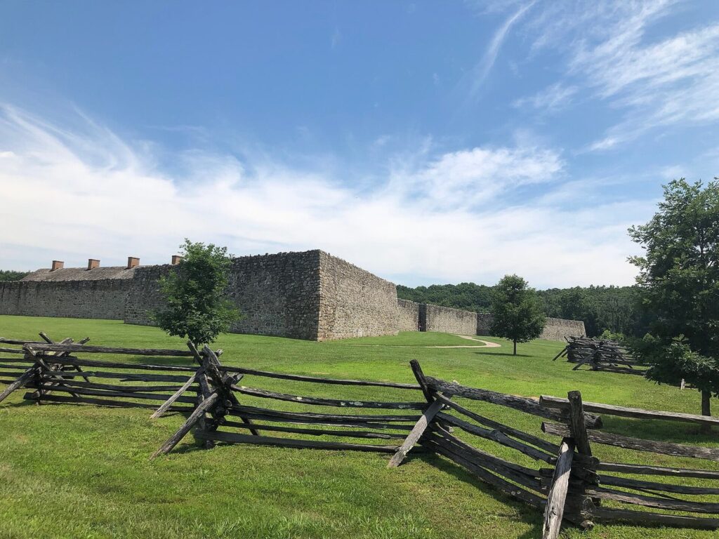 The long gray stone walls of Fort Frederick behind a split-rail fence on a mown lawn