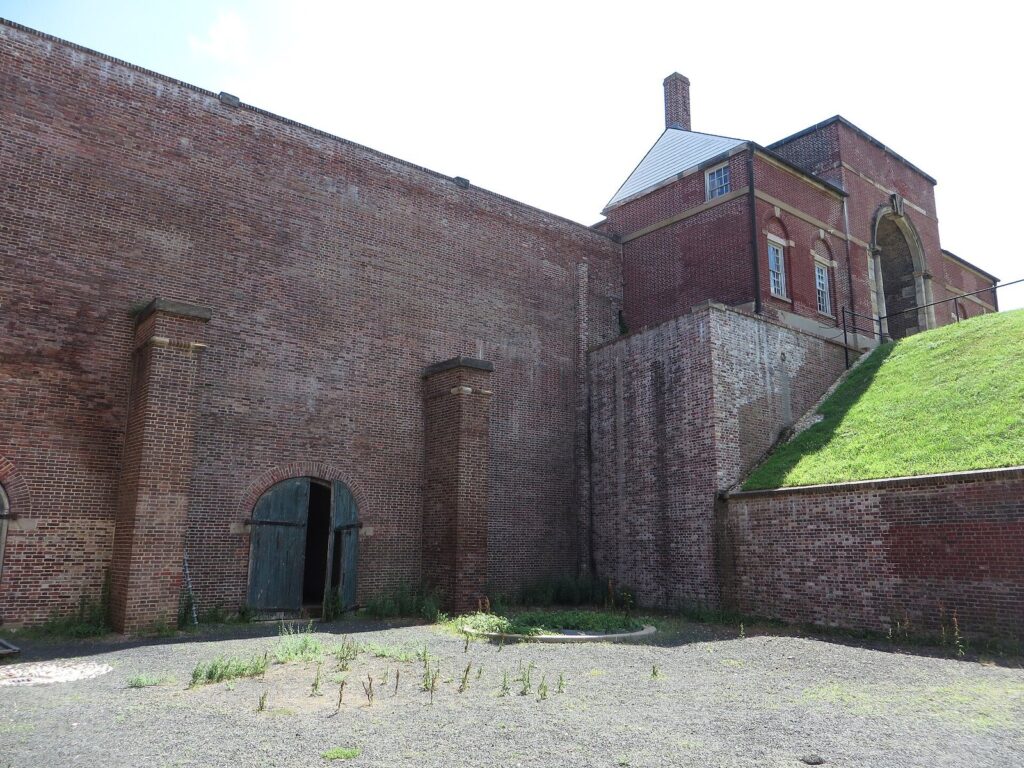 Tall weathered red-brick fort walls with a grassy earthwork slope and a brick building with an arched gateway