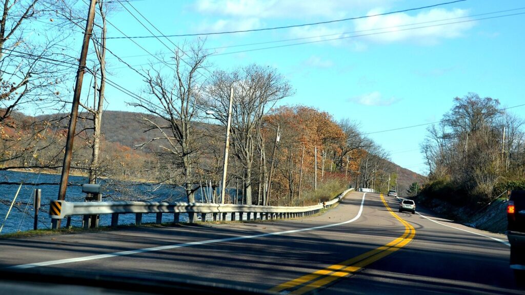 A two-lane country road curving beside the water under autumn trees and a blue sky