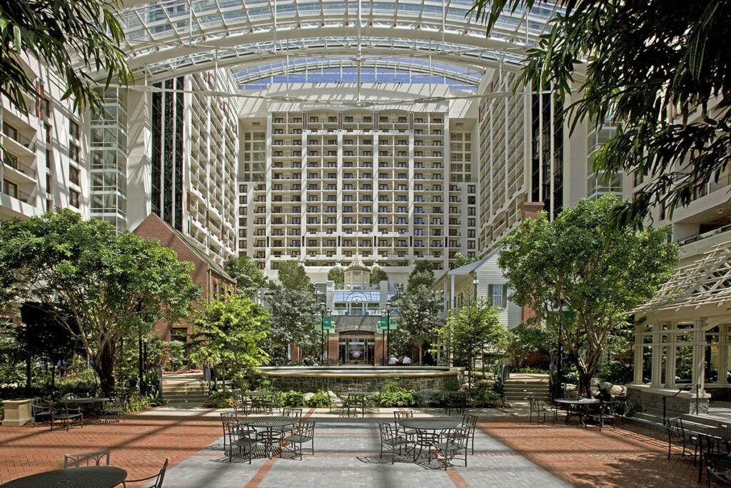 Tall glass-roofed hotel atrium with balconies on every floor, leafy trees, brick paving and cafe tables below