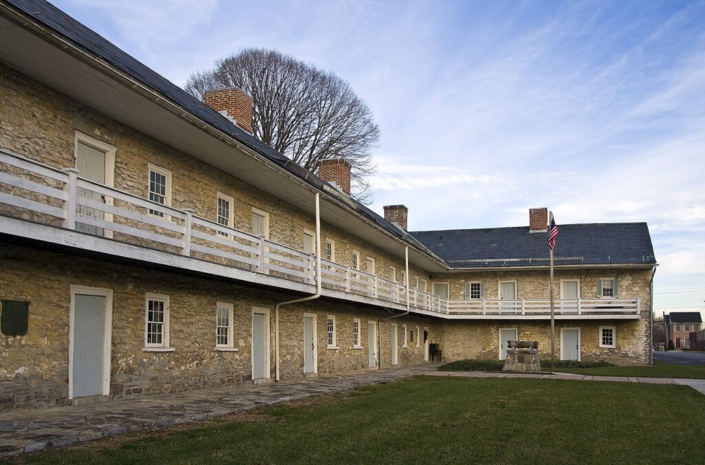 A long two-story fieldstone barracks with a white wooden gallery and a green lawn in front