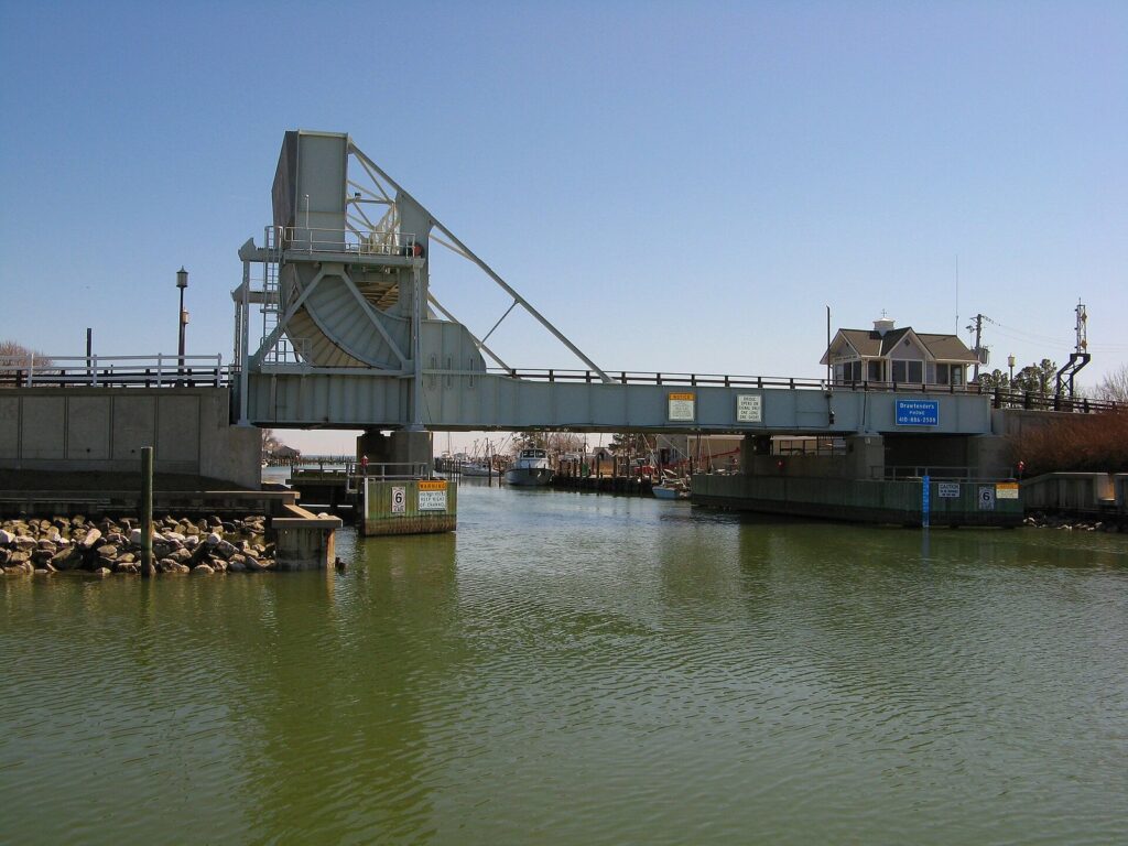 A steel lift drawbridge over a narrow channel with a small operator's house and workboats beyond