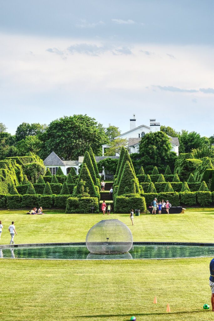 Clipped topiary hedges and pyramids around a lawn and a round pool, with a white house beyond