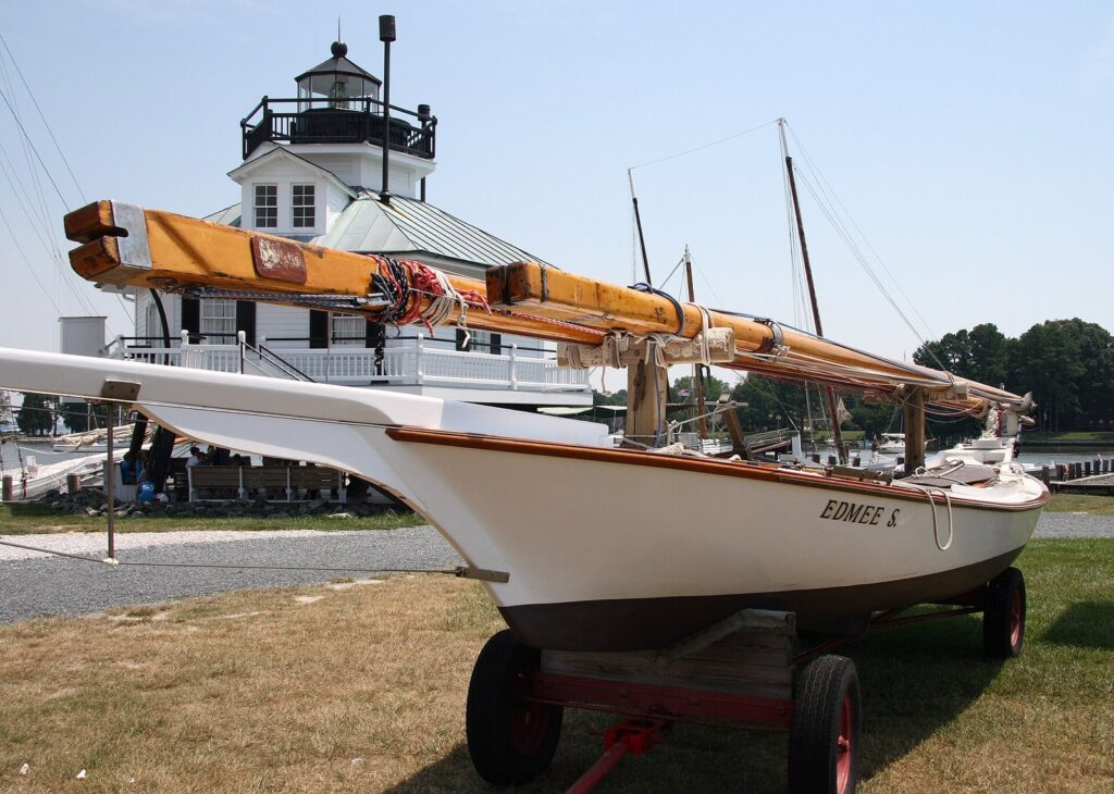 A sleek white sailing log canoe on a trailer on the grass, with the lighthouse behind it