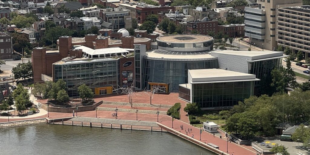 An aerial view of the Maryland Science Center's buildings beside the harbor promenade, with rowhouses behind