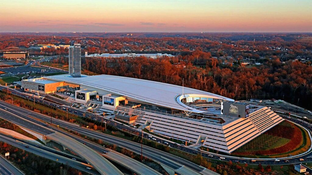 Aerial view of a casino resort with a tall hotel tower and a long, low building with a sloped glass front, surrounded by highway ramps and autumn trees
