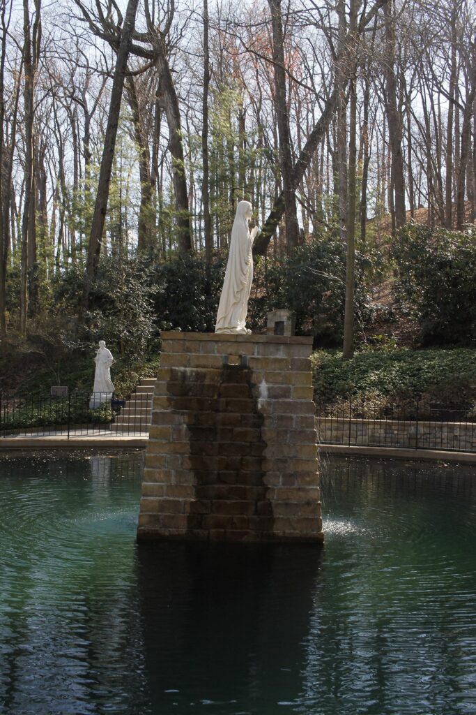 A white statue on a stone pedestal rising from a pond in a wooded garden