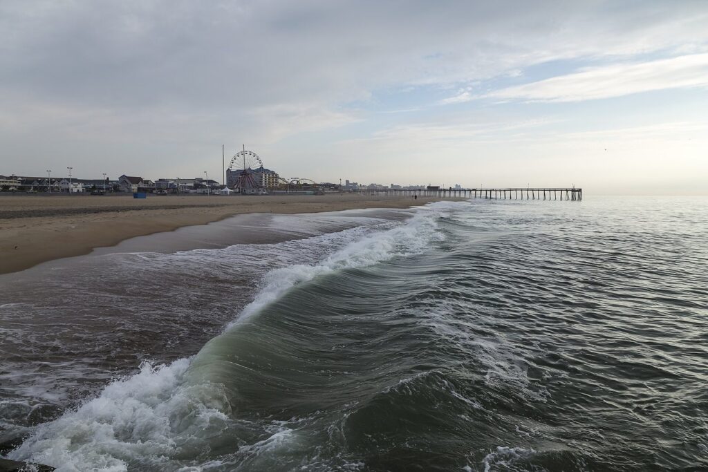 Waves washing up an almost empty beach toward the fishing pier and a Ferris wheel on an overcast morning