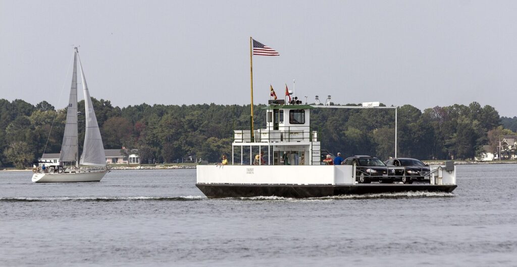A small open car ferry crossing a wide river beside a sailboat, with a wooded shore behind