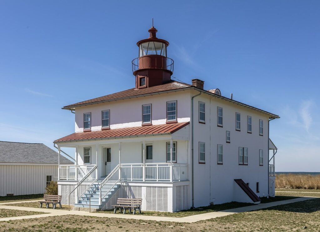 A white lighthouse house with a red roof and lantern under a blue sky, with dry grasses in front