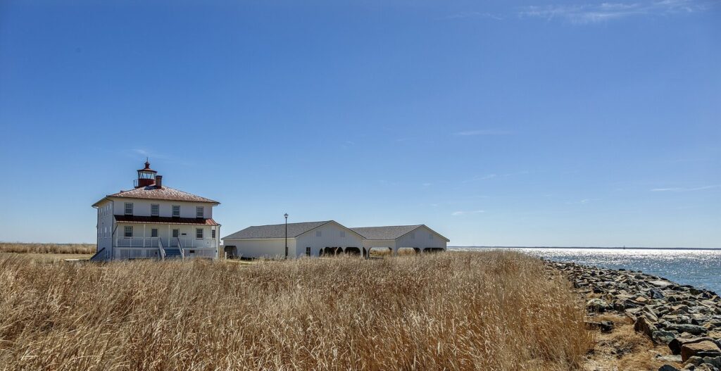 Point Lookout Lighthouse and a low white building behind tall marsh grass, with the Bay and a rock seawall to the right