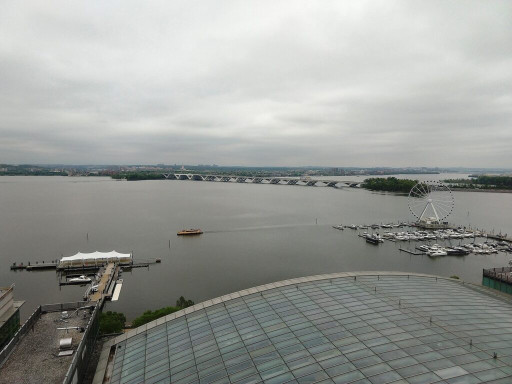View over the Potomac River from a hotel roof, with a covered pier, a Ferris wheel beside a marina, a yellow water taxi and a long arched highway bridge