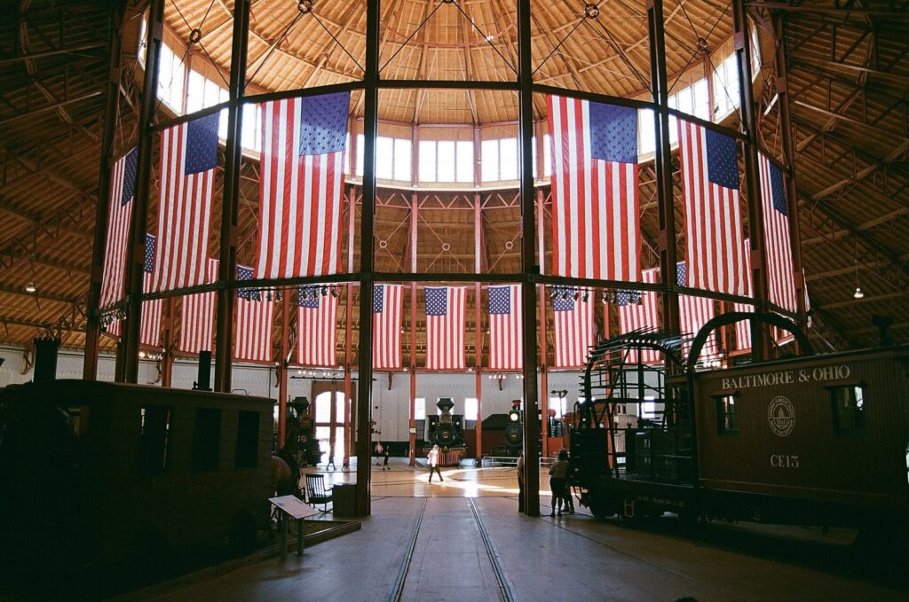 Historic steam locomotives beneath the wooden roof and tall windows of a round engine house hung with American flags