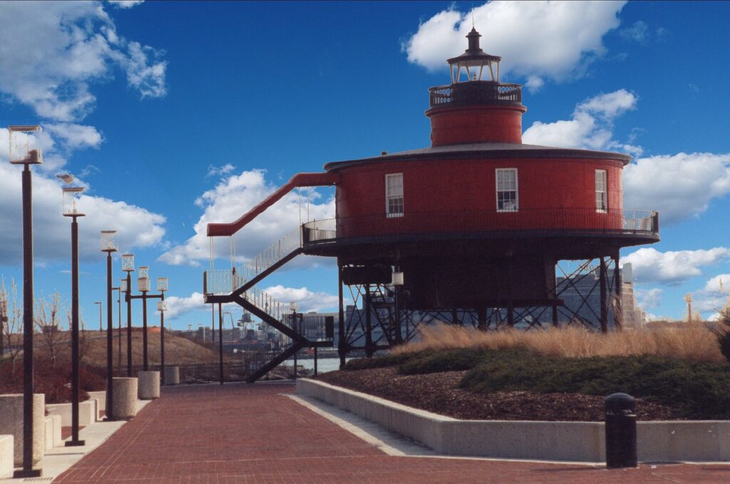 A round red screwpile lighthouse on iron legs beside the brick promenade under a blue sky
