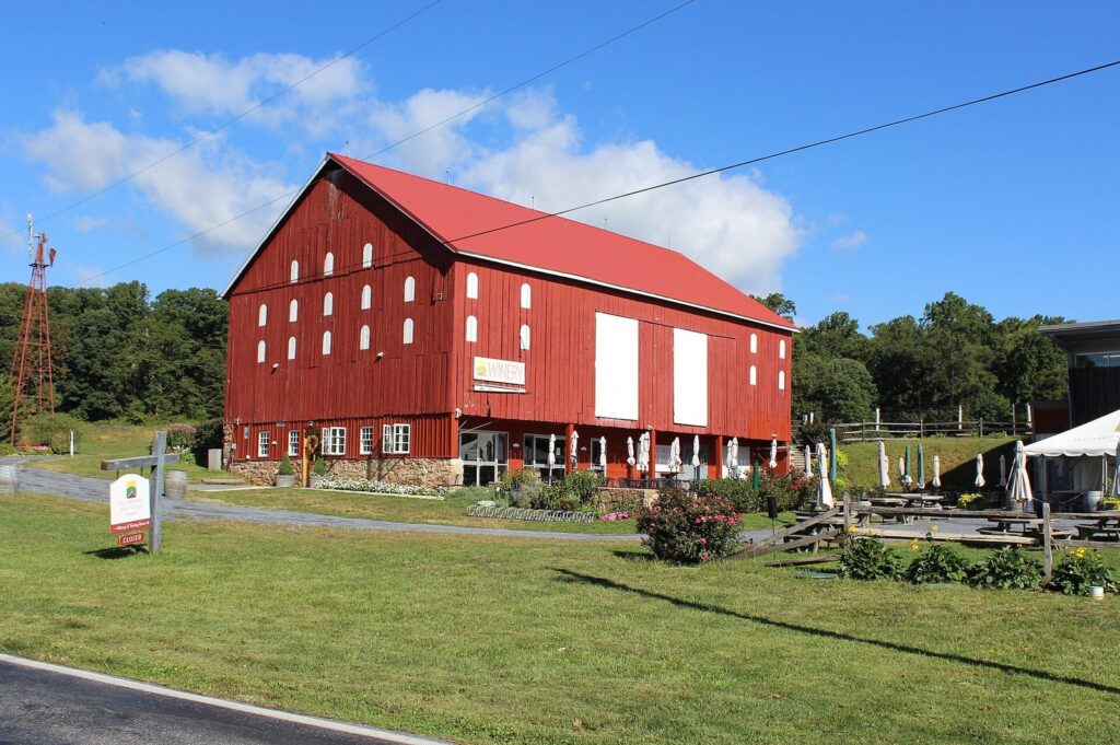 A big red bank barn with a winery sign, a stone foundation, patio umbrellas and a wide lawn under a blue sky