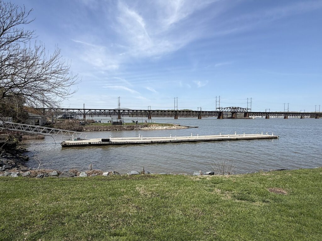 A long steel railroad bridge crossing a wide river beyond a floating dock and a grassy bank