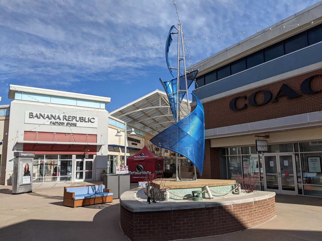 Outdoor shopping court with a blue metal sculpture, benches and outlet storefronts under a clear sky