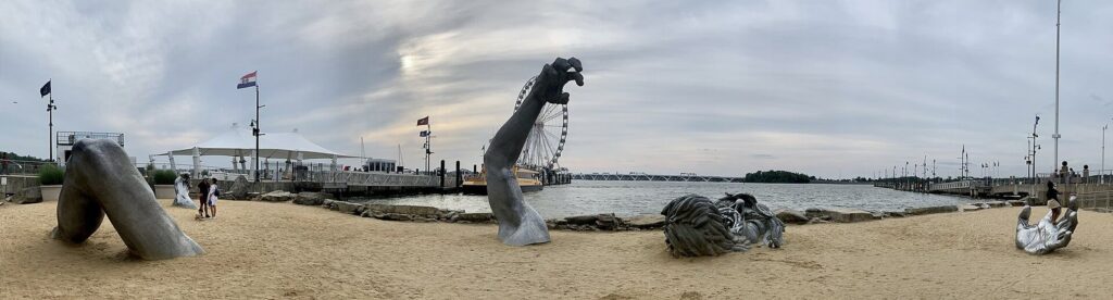 Panorama of a giant aluminum figure emerging from sand, with a raised hand, a head, a knee and a foot spread across a small beach by the river