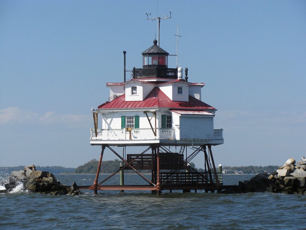 A white hexagonal screwpile lighthouse with a red roof standing on iron legs in open water