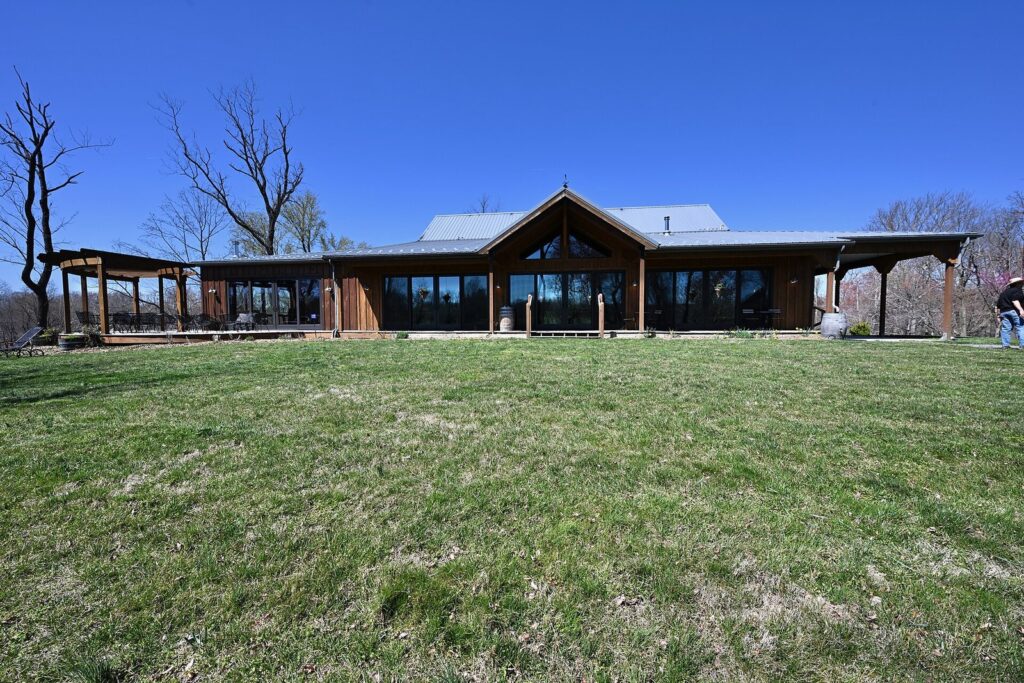 A long, low wood-and-glass tasting room with a deep porch across a wide green lawn under a clear blue sky