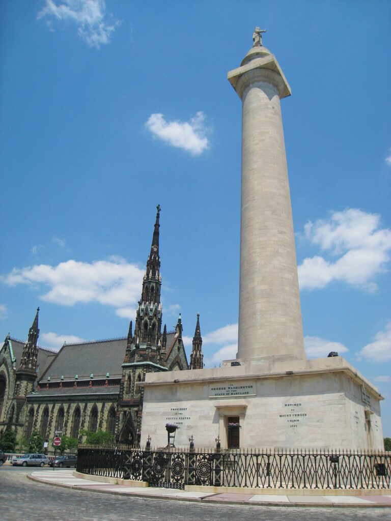 A tall white marble column topped by a statue, beside a Gothic church spire under a blue sky
