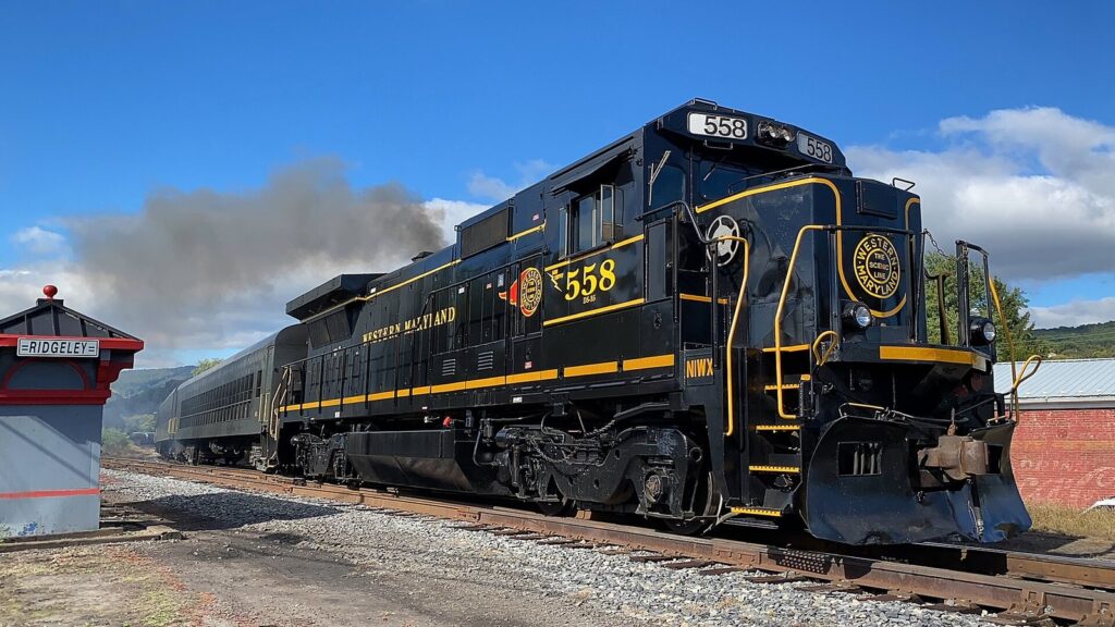 A black Western Maryland Scenic Railroad locomotive with passenger cars behind it beside a small depot under blue sky