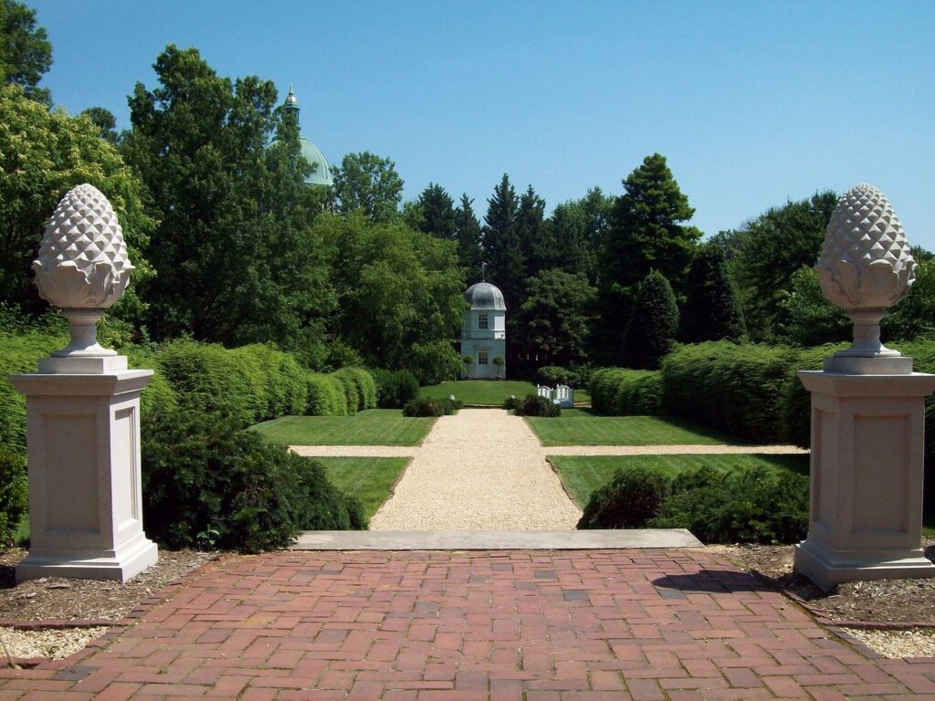 A formal garden path between stone pineapple finials, leading to a small white summerhouse among trees